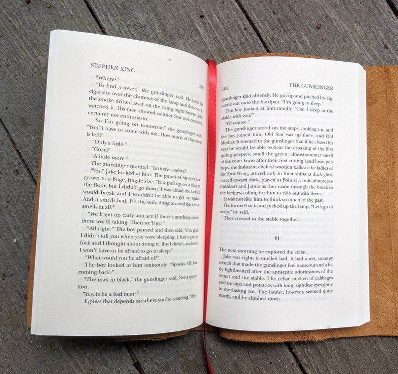 May include: Open book with a brown leather cover. The pages are open to a chapter titled "The Gunslinger" from the book "The Gunslinger" by Stephen King. The text on the page describes a scene where a gunslinger and a young boy are talking about a cellar.