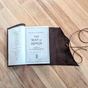 May include: A brown leather book cover with a red ribbon bookmark. The book is open to a page with the title "The Way of Kings" by Brandon Sanderson. The book is part of the Stormlight Archive series.
