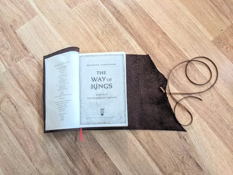 May include: A brown leather book cover with a red ribbon bookmark. The book is open to a page with the title "The Way of Kings" by Brandon Sanderson. The book is part of the Stormlight Archive series.