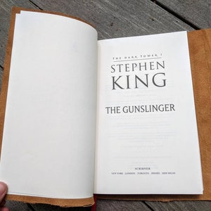May include: A brown leather-bound book with the title "The Gunslinger" by Stephen King. The book is part of the "The Dark Tower" series.