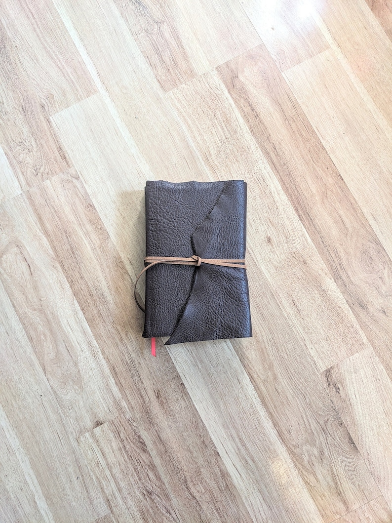 May include: A brown leather-bound journal with a leather strap closure. The journal is closed and lying flat on a light wood surface.