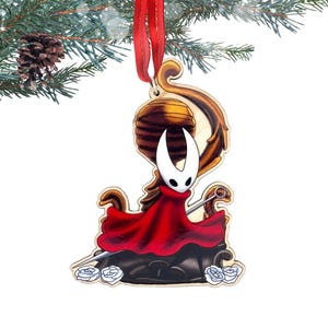 May include: Wooden Christmas ornament featuring a character in a red cloak and white mask. The ornament has a red ribbon for hanging and is decorated with a stylized design. The ornament is hanging from a pine branch.
