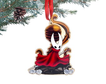 May include: Wooden Christmas ornament featuring a character in a red cloak and white mask. The ornament has a red ribbon for hanging and is decorated with a stylized design. The ornament is hanging from a pine branch.