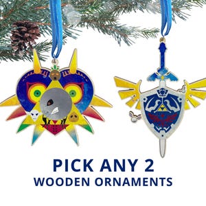 May include: Two wooden Christmas ornaments with blue ribbons hanging from a pine branch. One ornament is a colorful mask, the other is a sword and shield design. Text at the bottom reads "PICK ANY 2 WOODEN ORNAMENTS."