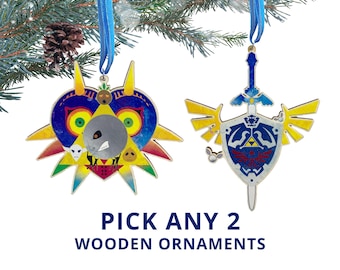 May include: Two wooden Christmas ornaments with blue ribbons hanging from a pine branch. One ornament is a colorful mask, the other is a sword and shield design. Text at the bottom reads "PICK ANY 2 WOODEN ORNAMENTS."