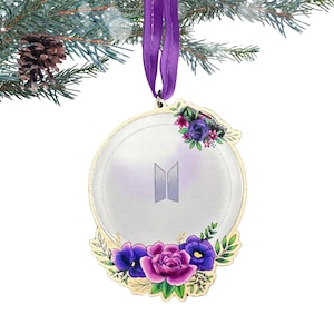 May include: A gold-colored Christmas ornament with a white background and the letters "BTS" in purple. The ornament is decorated with purple, pink, and green flowers.