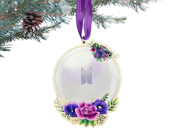 May include: A gold-colored Christmas ornament with a white background and the letters "BTS" in purple. The ornament is decorated with purple, pink, and green flowers.