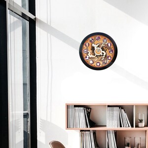 Pu&ograve; includere: Un orologio da parete rotondo con cornice nera. Il quadrante presenta un design a tema caff&egrave; con accenti marroni, arancioni e viola. L'orologio &egrave; appeso a una parete bianca.