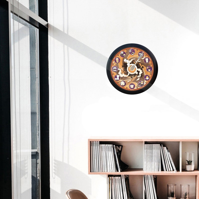 Pu&ograve; includere: Un orologio da parete rotondo con cornice nera. Il quadrante presenta un design a tema caff&egrave; con accenti marroni, arancioni e viola. L'orologio &egrave; appeso a una parete bianca.