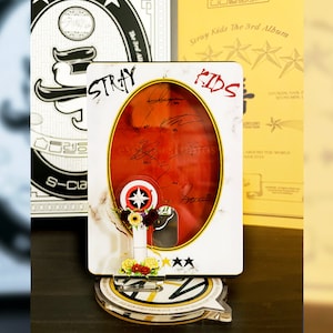 May include: A decorative stand featuring a red and white design with the text "STRAY KIDS" and signatures. The stand has a clear base with a logo and is adorned with flowers and stars. Other album covers are visible in the background.