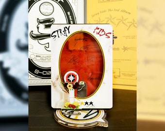 May include: A decorative stand featuring a red and white design with the text "STRAY KIDS" and signatures. The stand has a clear base with a logo and is adorned with flowers and stars. Other album covers are visible in the background.