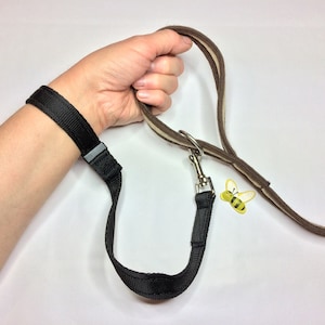 May include: A black wrist strap leash with a brown and beige leash attached to a silver clip. The leash is looped around a hand.