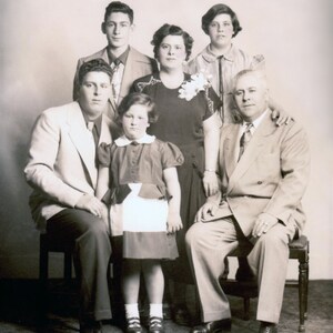 May include: A black and white photograph of a family of six. The family are standing in front of a plain background. The family are dressed in formal attire. The family are smiling.