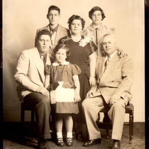 May include: A black and white photograph of a family of six. The family are standing in front of a white wall. The father is wearing a suit and tie. The mother is wearing a dress. The children are wearing dresses and suits. The family are smiling.