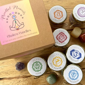 Puede incluir: Conjunto de siete pequeños frascos de vidrio con tapas, cada uno con una vela de soja. Los frascos están etiquetados con los nombres de los siete chakras: Raíz, Sacro, Plexo Solar, Corazón, Garganta, Tercer Ojo y Corona. Cada frasco también tiene un símbolo de color que representa el chakra. Los frascos están dispuestos sobre una superficie de madera con pequeñas piedras de varios colores.