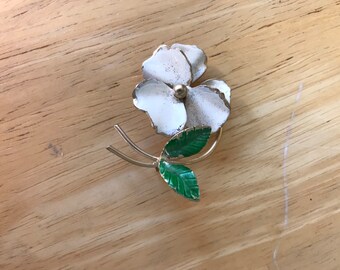 May include: A white enamel flower brooch with green enamel leaves and a gold tone finish.
