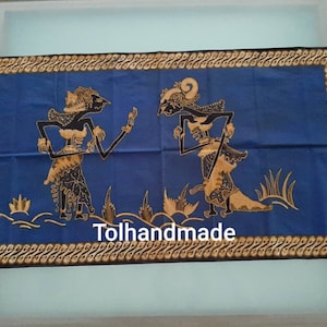 Puede incluir: Un tapiz de pared de tela azul con un borde dorado que presenta dos figuras negras en un estilo tradicional indonesio. Las figuras están representadas en una pose de baile y la tela tiene un diseño batik. El texto "Tolhandmade" está impreso en dorado en la parte inferior.