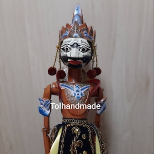 Peut inclure: Une marionnette en bois avec un visage peint et une coiffe élaborée. La marionnette porte une jupe noire et dorée avec des rubans jaunes. La marionnette est faite à la main et porte le texte "Tolhandmade" sur la poitrine.