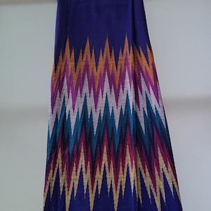 以下が含まれることがあります： 金、紫、ティール、白の色合いの鮮やかなジグザグ模様が特徴の、長い濃い青色の織物。生地はドレープされており、カラフルなデザインが際立っています。この織物は、スカーフまたは装飾品である可能性があります。