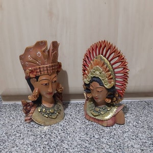 Peut inclure: Deux sculptures en bois sculptées de têtes de personnes avec des coiffures ornées. La sculpture de gauche a une coiffe brune avec des fleurs blanches et la sculpture de droite a une coiffe rouge avec des accents dorés.