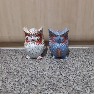 May include: Two hand-painted wooden owl figurines. The owl on the left is white with yellow, red, and green accents. The owl on the right is blue with red, yellow, and gold accents.