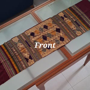May include: A burgundy and gold woven table runner with a geometric pattern. The runner is laid out on a glass table top.