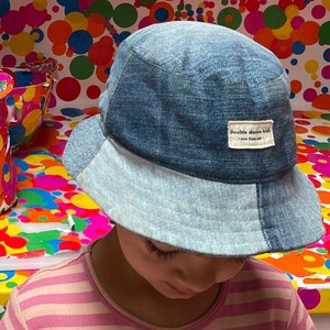May include: A child wearing a blue denim bucket hat with the text "double denim kids" on the front. The hat is sitting on top of the child's head, and the child is looking down.