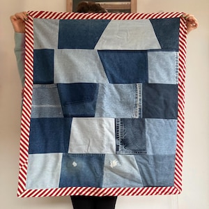 May include: A patchwork quilt made from repurposed denim jeans. The quilt features various shades of blue denim, arranged in a geometric pattern. The edges are finished with a red and white striped border.