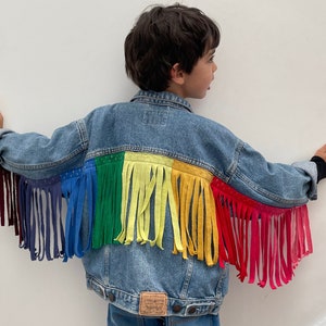 May include: A blue denim jacket with rainbow coloured fringe trim. The fringe is made of fabric strips sewn to the back of the jacket.