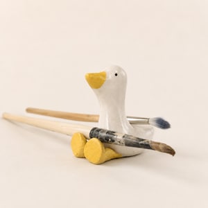 Könnte beinhalten: Eine weiße Keramik-Entenfigur mit gelbem Schnabel und gelben Füßen, die als Pinselablage dient. Die Ente ist so positioniert, dass zwei Pinsel auf ihrem Körper liegen, einer mit hellem Holzgriff und der andere mit einem dunklen Griff.