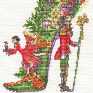 May include: Cross-stitch design of a Christmas stocking with a green base and purple trim, decorated with a fir tree, ornaments, and a Nutcracker soldier. A figure in a red coat and a Nutcracker doll are also present.