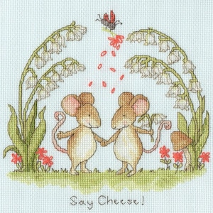May include: A cross-stitch pattern featuring two brown mice holding hands under an arch of white lily of the valley flowers. A ladybird flies above them, dropping red petals. Small red flowers and a mushroom are visible. Text reads: Say Cheese!