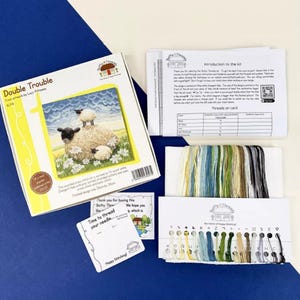 Pode incluir: Um kit de ponto cruz intitulado "Double Trouble" que apresenta uma ilustra&ccedil;&atilde;o colorida de duas ovelhas num campo de flores. O kit inclui um tecido estampado, fio de bordar e instru&ccedil;&otilde;es. O kit est&aacute; embalado numa caixa amarela e branca com uma imagem das ovelhas na parte da frente.