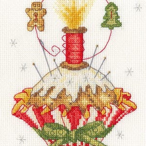 May include: Cross-stitch design featuring a festive Christmas ornament. The ornament is red and white with a green bow, adorned with a gingerbread man, Christmas tree, and star. Needles are placed around the ornament.