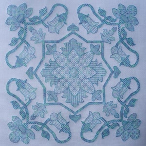 May include: A blue and green cross-stitch pattern with a floral design. The pattern features a central flower with eight petals and a repeating pattern of smaller flowers and leaves.