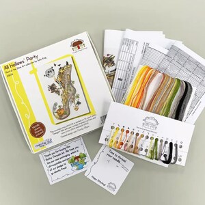 May include: A cross stitch kit featuring a whimsical illustration of a witch's boot with a cat, pumpkin, and other Halloween elements. The kit includes a printed pattern, embroidery floss, and a needle. The kit is packaged in a yellow box with the title "All Hallows' Party".