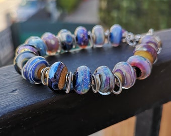 May include: A handcrafted bracelet featuring iridescent glass beads in shades of blue, purple, and gold. The beads are strung together with silver-colored metal wire. The bracelet is displayed on a black surface with a blurred background.