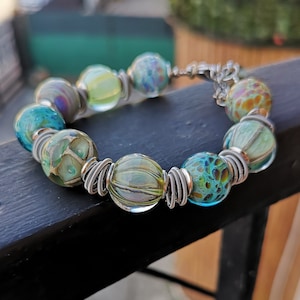 Puede incluir: Un brazalete hecho con cuentas de vidrio de colores y alambre plateado. Las cuentas son una mezcla de verde, azul y blanco con varios patrones.