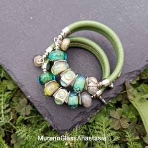 Puede incluir: Dos pulseras de cuero verde con cierres plateados y una fila de cuentas de vidrio de colores. Las cuentas son verdes, azules, blancas y amarillas.