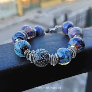 Könnte beinhalten: Ein Armband aus blauen und violetten Glasperlen mit silbernen Draht-Akzenten. Die Perlen haben eine Vielzahl von Texturen und Mustern, darunter Wirbel, Flecken und eine strukturierte Kugel.