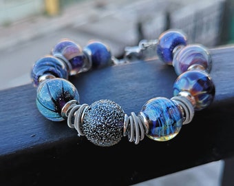 Op de afbeelding: Een armband gemaakt van blauwe en paarse glazen kralen met zilveren draadaccenten. De kralen hebben een verscheidenheid aan texturen en patronen, waaronder swirls, stippen en een textuurbol.