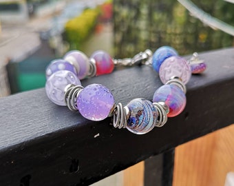 May include: A purple, pink, and blue glass bead bracelet with silver wire accents. The beads are round and have a textured surface.