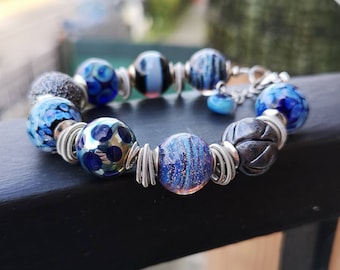 May include: A blue and silver bracelet with a variety of glass beads, including some with a speckled pattern. The bracelet is made with wire and metal accents.