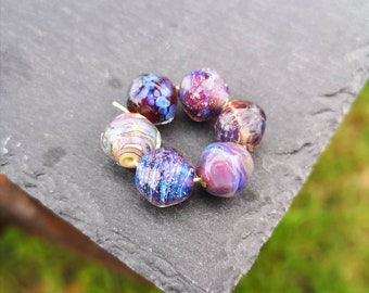 Op de afbeelding: Zes kleurrijke glazen kralen met swirls van blauw, paars en goud. De kralen zijn in een cirkel op een donkergrijs oppervlak gerangschikt.