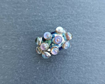 Puede incluir: Una cuenta de vidrio multicolor con un patrón azul, verde y blanco. La cuenta está cubierta de pequeños círculos iridiscentes.