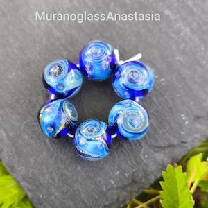 May include: A cluster of six round, blue glass beads with swirling patterns and silver accents, strung together on a white cord. The beads are arranged in a circular shape, with the text "MuranoGlassAnastasia" at the top.