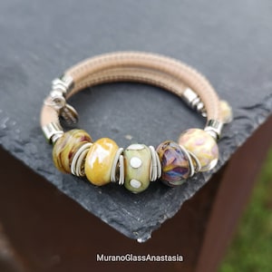 May include: A tan leather bracelet with silver accents and five colorful glass beads. The beads are yellow, green, white, purple, and orange.