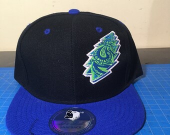 May include: A black and blue baseball cap with a green and white embroidered patch of a stylized pine tree. The cap has a blue brim and a white label that says "Pit Bull Snapback High Quality Premium Headwear".
