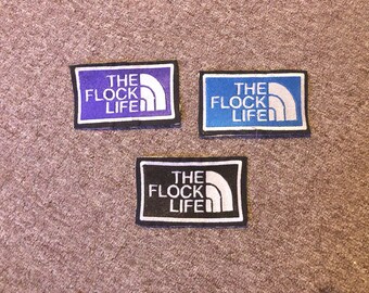 May include: Three embroidered patches with the text "The Flock Life" in white lettering on a black, blue, and purple background. Each patch features a white mountain range graphic.