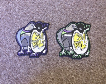 May include: Two black and white penguin cartoon patches with green and purple accents. The penguins are hugging and have a yellow oval with the text "Be! Real!" on it.